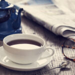An outdoor table is set with a teapot, teacup, newspaper, and glasses.