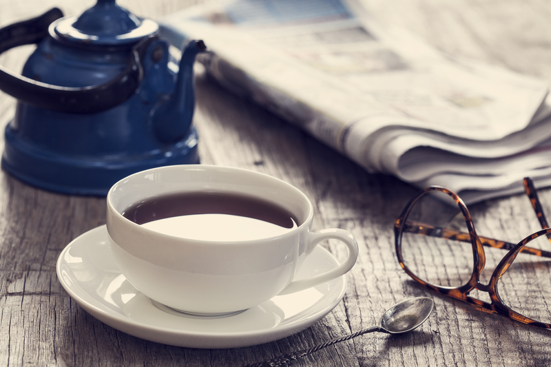 An outdoor table is set with a teapot, teacup, newspaper, and glasses.